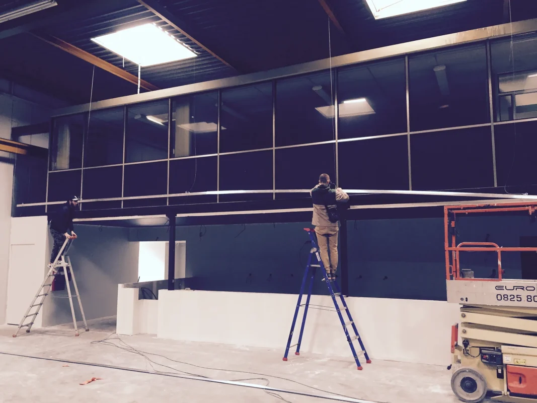 Deux techniciens posent des éléments vitrés sous une mezzanine, sur échelles, dans un hall en travaux.