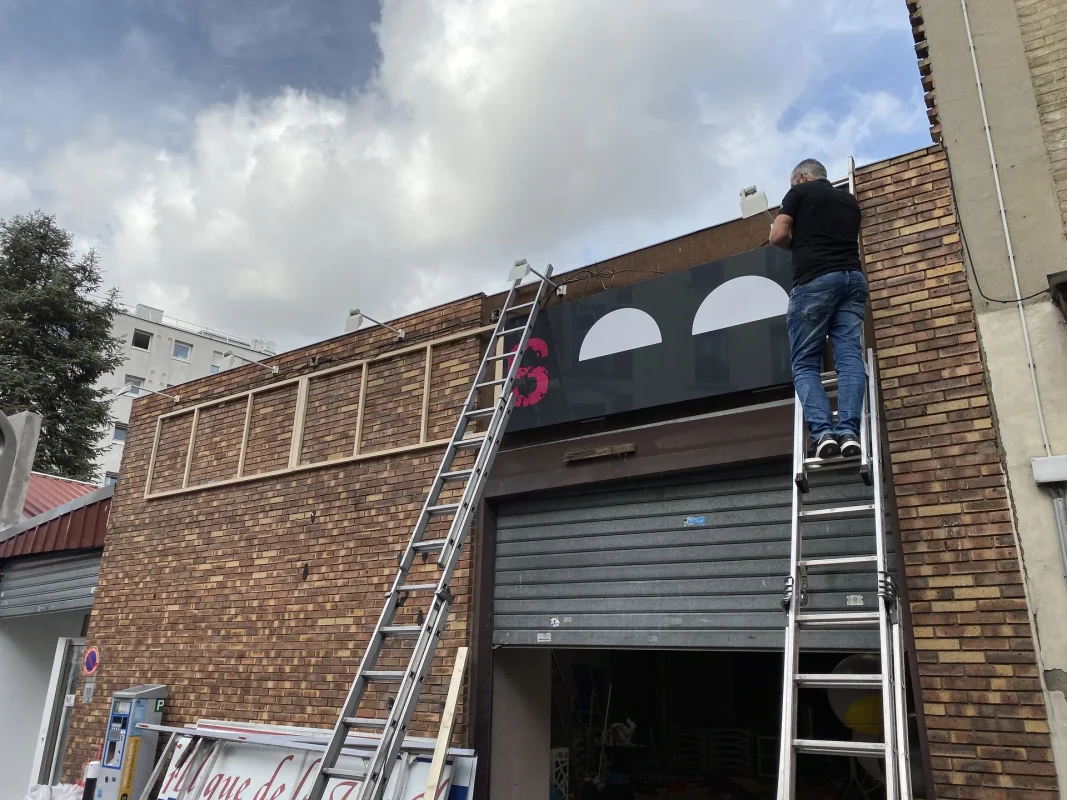 Technicien sur échelle posant un grand panneau noir sur une façade en briques.