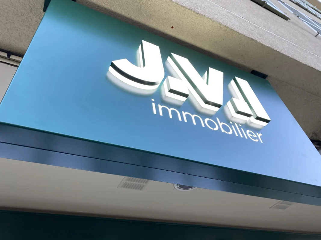Caisson lumineux JVL immobilier éclairé, lettres blanches sur panneau bleu-vert.