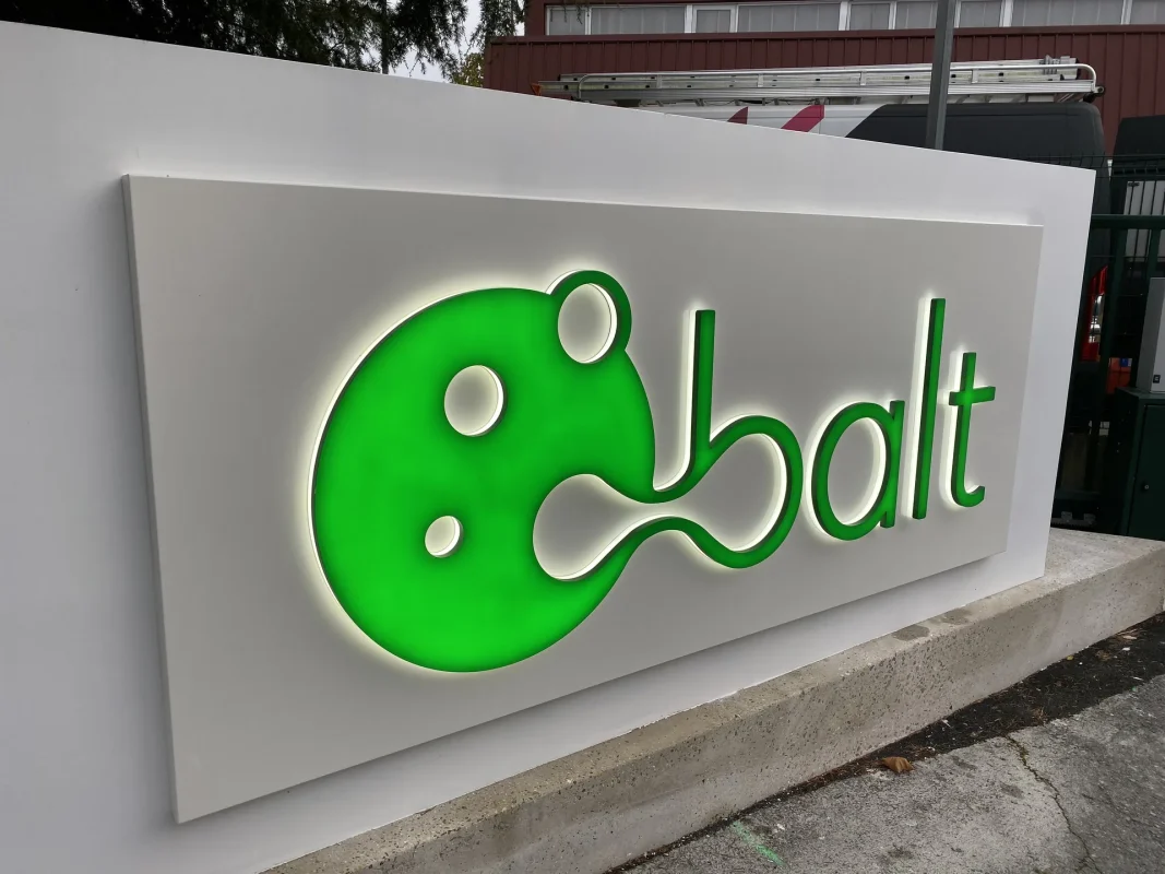 Enseigne balt en lettres vertes rétroéclairées sur panneau blanc, visible de côté, sur un mur d'entrée.