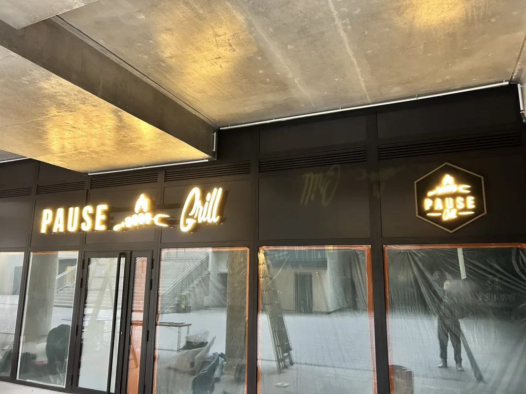 Lettres Pause Grill éclairées et hexagone lumineux sur une façade sombre, sous une structure en béton.