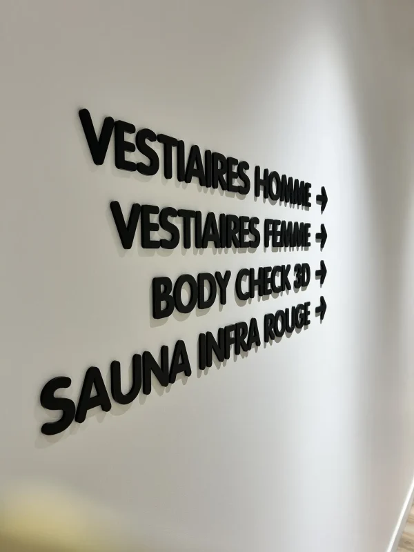 Lettres noires en relief indiquant vestiaires homme et femme, body check 3D, sauna infrarouge, avec flèches.