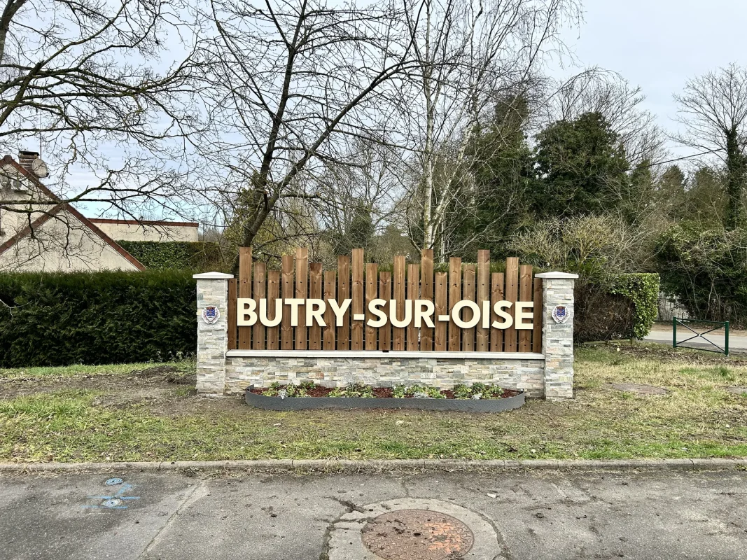 Panneau d'entrée de Butry-sur-Oise : lettres bois sur clôture en lames, muret en pierre et massifs.