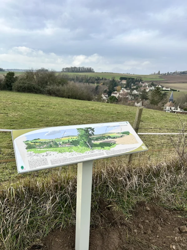 Panneau pupitre à illustration de paysage sur un poteau bois, dans un champ, avec un village au loin.